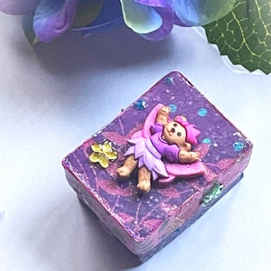 Puede incluir: Una pequeña caja púrpura con un acabado brillante y una figura de hada en la tapa. El hada lleva un vestido rosa y una corona. La caja está decorada con flores y purpurina.