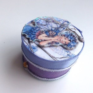 Puede incluir: Una pequeña caja redonda de color morado y blanco con una ilustración de hada azul y blanca en la tapa. La caja tiene un borde plateado y un botón dorado.