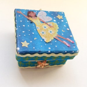 Puede incluir: Una pequeña caja azul con un diseño de hada amarilla y blanca. La caja tiene un fondo blanco con puntos blancos y estrellas. El hada lleva un vestido amarillo con flores blancas y tiene un halo blanco. La caja tiene un borde verde y amarillo alrededor de la parte superior.
