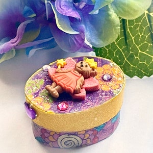 Puede incluir: Una pequeña caja redonda de purpurina morada y amarilla con una figurita de oso hada rosa y naranja en la parte superior. La caja tiene un patrón floral y detalles de purpurina morada.