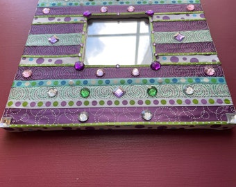 May include: A purple, green, and white striped mirror frame with rhinestones and a small mirror in the center.