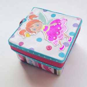 Puede incluir: Una pequeña caja cuadrada con un diseño de lunares coloridos. La caja presenta un hada con alas rosadas y un vestido rosa. El hada está rodeada de lunares coloridos.