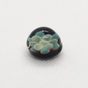 May include: A small, round glass bead with a black background and a white and blue flower design.