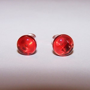 Puede incluir: Dos tapones para los oídos redondos rojos con un poste transparente. Los tapones para los oídos están hechos de un material translúcido y están colocados sobre un fondo blanco. Los tapones para los oídos son de un color rojo vibrante y sólido.