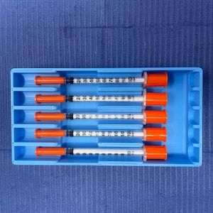 May include: A blue tray holds five syringes with orange caps and clear barrels. Each syringe has black measurement markings from 0 to 100. The tray is on a blue textured surface.