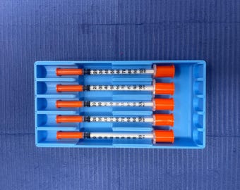 May include: A blue tray holds five syringes with orange caps and clear barrels. Each syringe has black measurement markings from 0 to 100. The tray is on a blue textured surface.