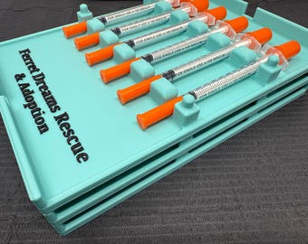 Puede incluir: Una bandeja organizadora de color turquesa con varias jeringas, cada una con una tapa naranja. La bandeja tiene el texto "Ferret Dreams Rescue & Adoption" impreso. Las jeringas están etiquetadas como "FOR SINGLE USE U-100 INSULIN".