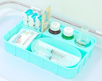 Puede incluir: Un organizador de plástico azul claro con múltiples compartimentos para suministros médicos. Incluye una botella transparente, una botella verde, una botella blanca, un paquete de gasas, un paquete de bastoncillos de algodón, un paquete de jeringas y un paquete de guantes médicos.
