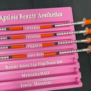 May include: A pink plastic organizer labeled "Ageless Beauty Aesthetics" holds several syringes with orange plungers. The organizer has slots labeled for various cosmetic procedures, including "Frontalis" and "Crows feet."
