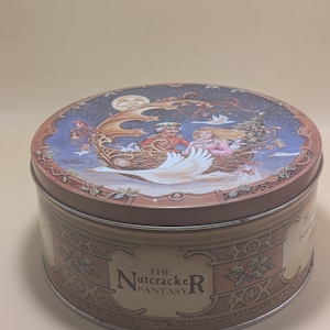 May include: A circular metal tin with a lid displaying a vibrant illustration from "The Nutcracker Fantasy." The lid shows a scene with figures in a boat pulled by a swan under a moonlit sky. The tin's side has the text "The Nutcracker Fantasy."