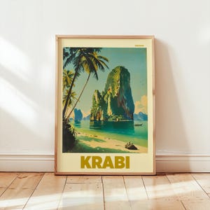 May include: Framed art print depicting a tropical beach scene with palm trees, turquoise water, and a large rock formation. The word "KRABI" is printed in gold block letters at the bottom of the print. The print is in a wooden frame.