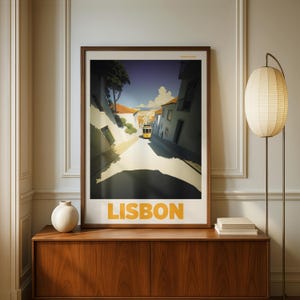 Puede incluir: Una impresión artística enmarcada que presenta una escena callejera en Lisboa, Portugal, con un tranvía amarillo en una calle soleada. La palabra "LISBON" está impresa en letras grandes de color naranja en la parte inferior. La impresión se exhibe en un mueble de madera.