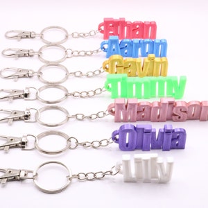 May include: A collection of personalized keychains, each featuring a different name in a 3D-printed style. The names are in various colors, including pink, blue, yellow, green, mauve, purple, and white. Each keychain has a silver clasp and chain.