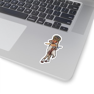 Op de afbeelding: Een sticker met een gestileerde illustratie van een krijger met een donkere huid, gekleed in een witte en bruine outfit, met twee gebogen messen. De vrouw heeft donker haar en gouden accenten. De sticker staat op een laptop.