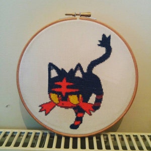May include: A cross-stitch embroidery of a red and black Pokemon character, with a white background. The character is a cat-like creature with a red star on its forehead and yellow eyes.