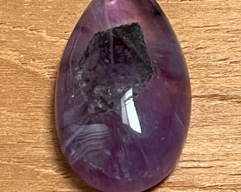 May include: A polished, teardrop-shaped amethyst gemstone with a dark, textured inclusion. The stone displays a gradient of purple hues, from deep violet to translucent lavender. The gemstone is set against a wooden surface.