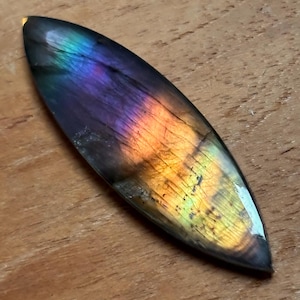 May include: A marquise-shaped labradorite gemstone cabochon featuring a polished surface with iridescent flashes of purple, blue, and golden-yellow light. The stone displays natural internal fractures and a smooth, reflective finish throughout.