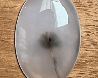 May include: Oval-shaped, translucent gemstone with a light gray hue and a central dark spot. The stone has a polished surface, reflecting light, and is set against a wooden background. The gemstone's shape and color suggest it could be used in jewelry.