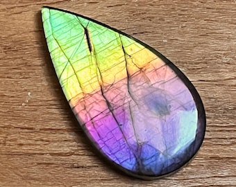 May include: A teardrop-shaped gemstone with a vibrant display of colors. The stone transitions from green to yellow, then to pink and purple. The surface has natural cracks and a polished finish, set against a wooden background.