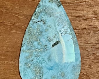 May include: A teardrop-shaped gemstone with a light blue and white marbled pattern. The polished surface reflects light, showcasing the stone's natural textures and variations. The stone is set against a wooden background.