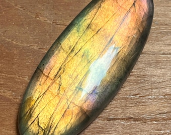 May include: An oval-shaped labradorite gemstone featuring a polished, smooth surface with visible internal fractures. The stone displays a vibrant play of color, transitioning from deep charcoal and green tones to bright golden-yellow and pink hues.