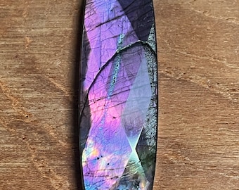 May include: A rectangular, polished gemstone with a faceted surface. The stone displays a vibrant play of color, with hues of purple, pink, and blue. The gemstone is set against a wooden background.