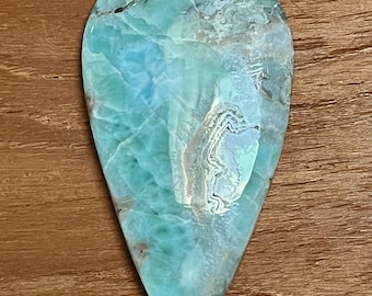 May include: A teardrop-shaped gemstone with a light blue and white marbled pattern. The polished stone has a smooth surface and is set against a wooden background. The gemstone is likely used for jewelry or decorative purposes.