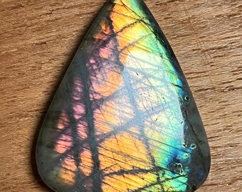 May include: A polished, teardrop-shaped labradorite gemstone. The stone displays a vibrant play of color, with iridescent flashes of orange, yellow, green, and blue across a dark gray base. The gemstone is set against a wooden surface.