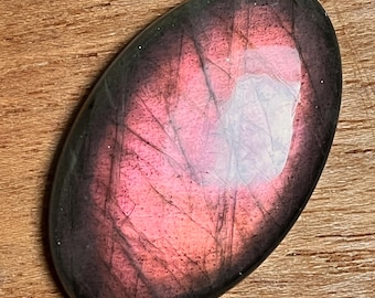 May include: An oval-shaped gemstone with a polished surface. The stone features a blend of pink and purple hues, with darker edges and lighter, reflective areas in the center. The gemstone is set against a wooden background.