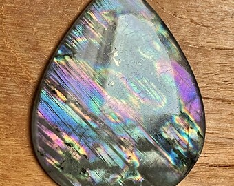 May include: A teardrop-shaped labradorite gemstone with iridescent colors of blue, purple, and green. The polished stone displays a play of light across its surface, set against a wooden background. The gemstone is a natural, unique piece.