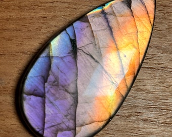 May include: A teardrop-shaped labradorite gemstone with a polished surface. The stone displays a vibrant color palette of purple, orange, and yellow hues, with dark veining throughout. The gemstone is set against a wooden background.