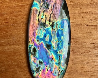May include: Oval-shaped labradorite gemstone with a polished surface. The stone displays iridescent colors of blue, purple, and green, with dark inclusions. The gemstone is set against a wooden background.