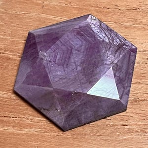 May include: A faceted, hexagonal amethyst crystal with a deep purple hue. The gemstone is cut with multiple flat surfaces, reflecting light and creating a geometric shape. The crystal sits on a wooden surface.