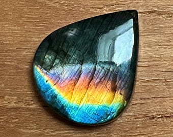 May include: A polished, teardrop-shaped labradorite gemstone. The stone displays a dark, iridescent surface with a vibrant rainbow of colors, including blue, yellow, and orange. The gemstone is set against a wooden background.