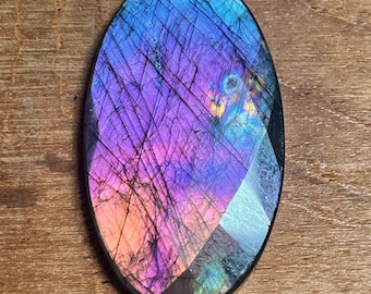 May include: Oval-shaped labradorite gemstone with a polished surface. The stone displays a vibrant play of color, with hues of blue, purple, and orange. The gemstone is set against a wooden background.