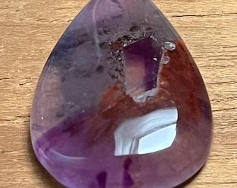 May include: A teardrop-shaped gemstone with a polished surface. The stone features a blend of purple, red, and clear hues, with internal inclusions. The gemstone is set against a wooden surface.