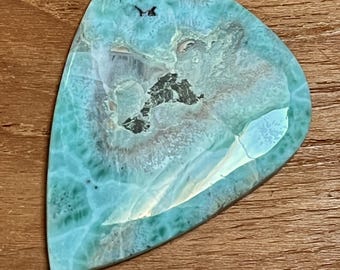 May include: A polished, teardrop-shaped gemstone with a striking turquoise and white pattern. The stone features dark gray inclusions and a glossy surface, set against a warm, wooden background. The gemstone's unique color and pattern make it a beautiful piece.