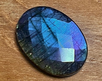 May include: Oval-shaped labradorite gemstone with a polished, faceted surface. The stone displays a vibrant play of color, with blues and purples dominating. The gemstone is set against a wooden background.