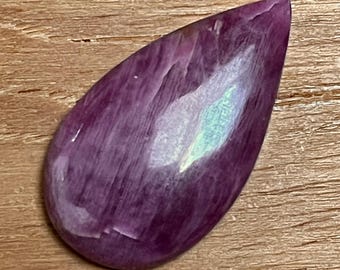 May include: A polished, teardrop-shaped gemstone in shades of deep purple with lighter streaks and a subtle iridescent sheen. The stone is set against a wooden surface.