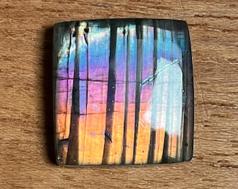 May include: A square-cut labradorite gemstone with a vibrant display of color. The stone features a mix of purple, blue, and orange hues, with vertical lines creating a striped effect. The gemstone is set against a wooden surface.