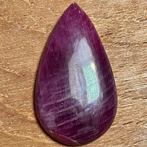 May include: A polished, teardrop-shaped gemstone with a deep magenta hue. The stone has a smooth, reflective surface with subtle internal striations. The gem is set against a warm, wooden background, highlighting its rich color and natural beauty.