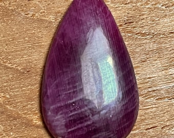May include: A polished, teardrop-shaped gemstone with a deep magenta hue. The stone has a smooth, reflective surface with subtle internal striations. The gem is set against a warm, wooden background, highlighting its rich color and natural beauty.