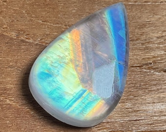 May include: A teardrop-shaped, polished gemstone with a pearlescent sheen. The stone displays a spectrum of colors, including blues, greens, and yellows, creating a rainbow effect. The gem is set against a wooden surface.