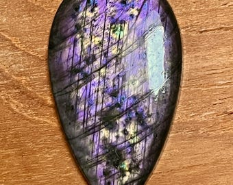 May include: A teardrop-shaped gemstone with a polished surface. The stone displays a vibrant play of color, with shades of purple, blue, and green. Dark lines and inclusions add texture to the iridescent surface. The gemstone is set against a wooden background.