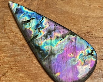 May include: A teardrop-shaped labradorite gemstone with iridescent colors of blue, purple, and green. The polished stone has a smooth surface and is set against a wooden background.
