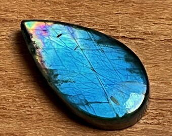 May include: A teardrop-shaped labradorite gemstone with a polished surface. The stone displays a vibrant blue color with iridescent flashes of green, purple, and yellow. Fine lines and natural inclusions are visible within the stone. The gemstone is set against a wooden background.