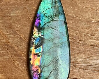 May include: A teardrop-shaped labradorite gemstone with iridescent colors of blue, green, and purple. The polished stone has a smooth surface with natural cracks and inclusions, set against a wooden background.