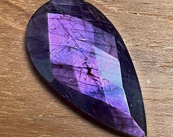 May include: A teardrop-shaped gemstone with a deep purple hue and internal crystalline patterns. The stone has a polished, faceted surface that reflects light, creating a shimmering effect. The gemstone is set against a wooden background.