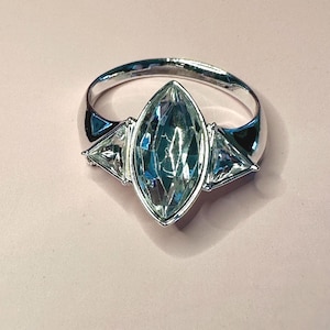 May include: A silver-toned ring featuring a large, central marquise-cut light blue gemstone. The center stone is flanked by two smaller, triangular-cut clear gemstones on either side, all set within a polished metallic band with a smooth finish.