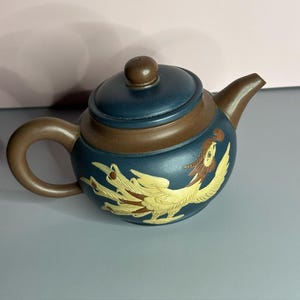May include: A small, ceramic teapot with a dark blue and brown color scheme. The teapot features a detailed, gold-colored phoenix design on its side. The lid and handle are brown, and the spout is curved.